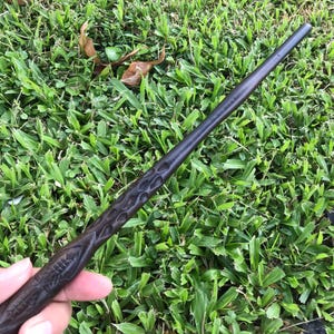 May include: A dark brown wooden wand with intricate carvings. The wand is held in a hand against a green grassy background.