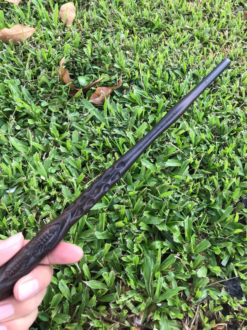 May include: A dark brown wooden wand with intricate carvings. The wand is held in a hand against a green grassy background.