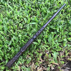 May include: A dark brown wooden wand with intricate carvings. The wand is lying on a bed of green grass.