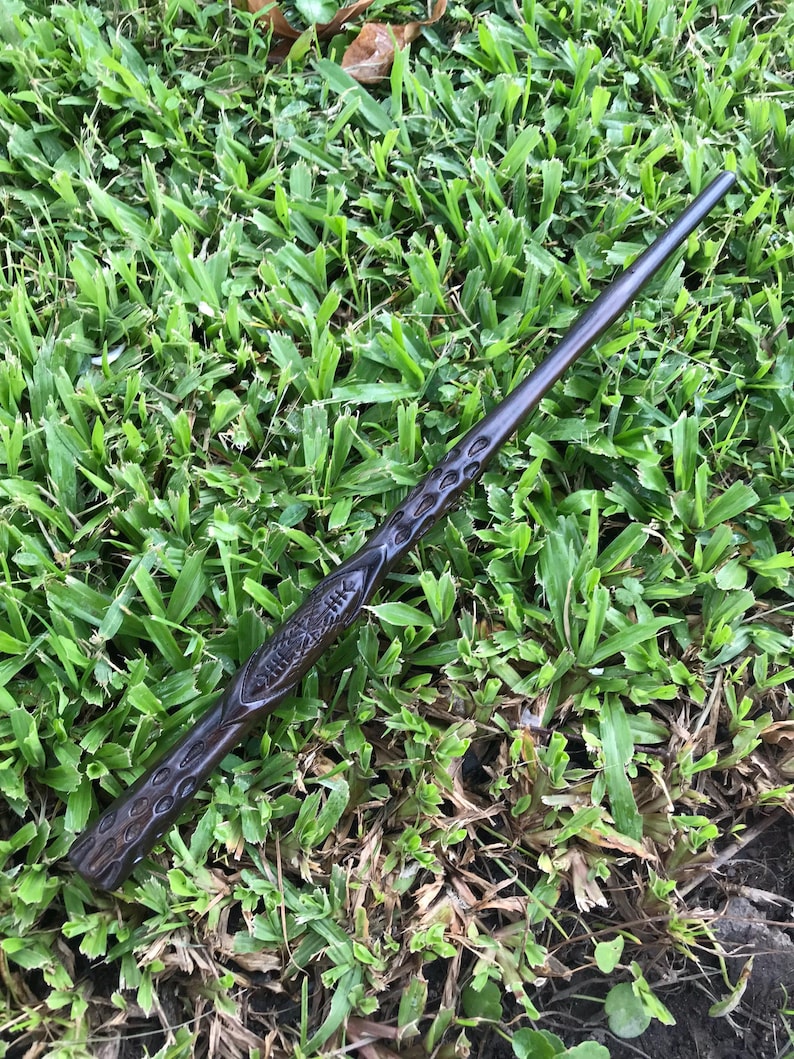 May include: A dark brown wooden wand with intricate carvings. The wand is lying on a bed of green grass.