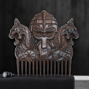 May include: A dark brown wooden comb with a Viking helmet design. The comb features intricate carvings of a helmeted face, two dragon-headed figures, and a series of teeth. The comb is displayed on a black surface.