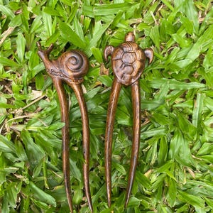 May include: Three wooden hair pins with animal carvings. The pins are brown and have a natural wood finish. The carvings are of a snail, a turtle, and a fish.