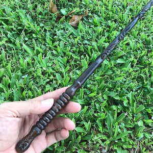 May include: A dark brown wooden wand with intricate carvings and a twisted handle. The wand is held in a hand against a green grassy background.