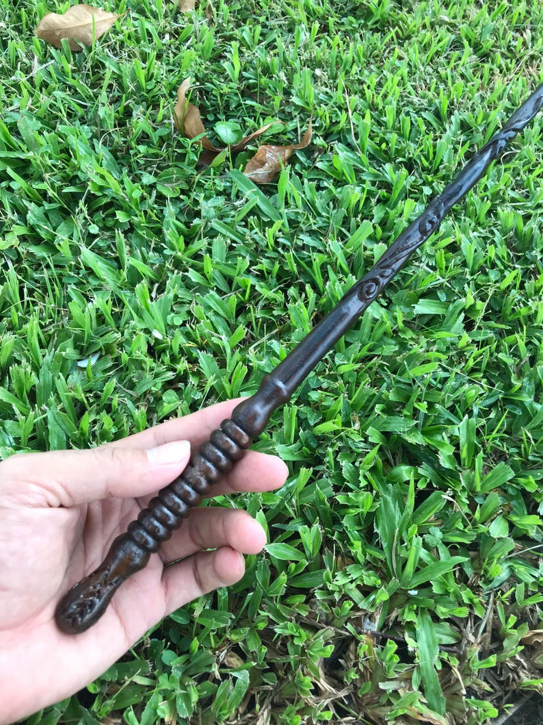May include: A dark brown wooden wand with intricate carvings and a twisted handle. The wand is held in a hand against a green grassy background.