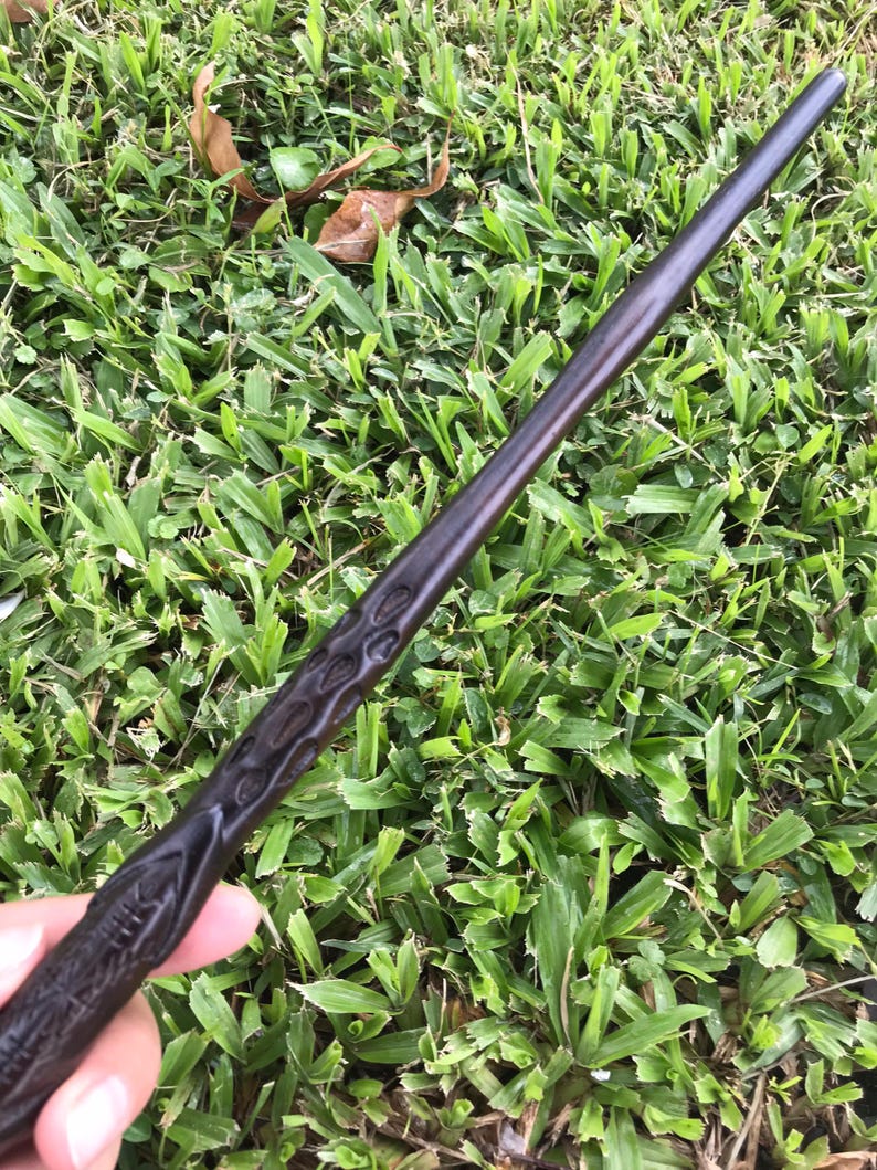 May include: A dark brown wooden wand with intricate carvings. The wand is approximately 33 cm long and has a pointed tip.