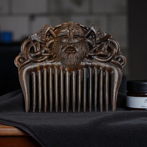 May include: A wooden comb with a carved Viking face and axes design. The comb is dark brown and sits on a black cloth next to a jar with a black lid and a white label.
