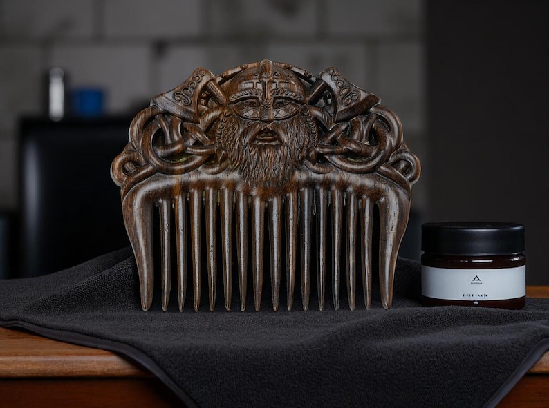 May include: A wooden comb with a carved Viking face and axes design. The comb is dark brown and sits on a black cloth next to a jar with a black lid and a white label.