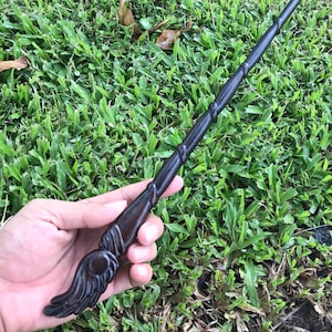 May include: A dark brown wooden wand with a carved, feathered design at the end. The wand is held in a hand against a background of green grass.