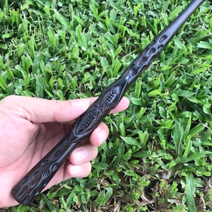 May include: A dark brown wooden wand with intricate carvings. The wand is held in a hand, with the tip of the wand pointing towards the right.
