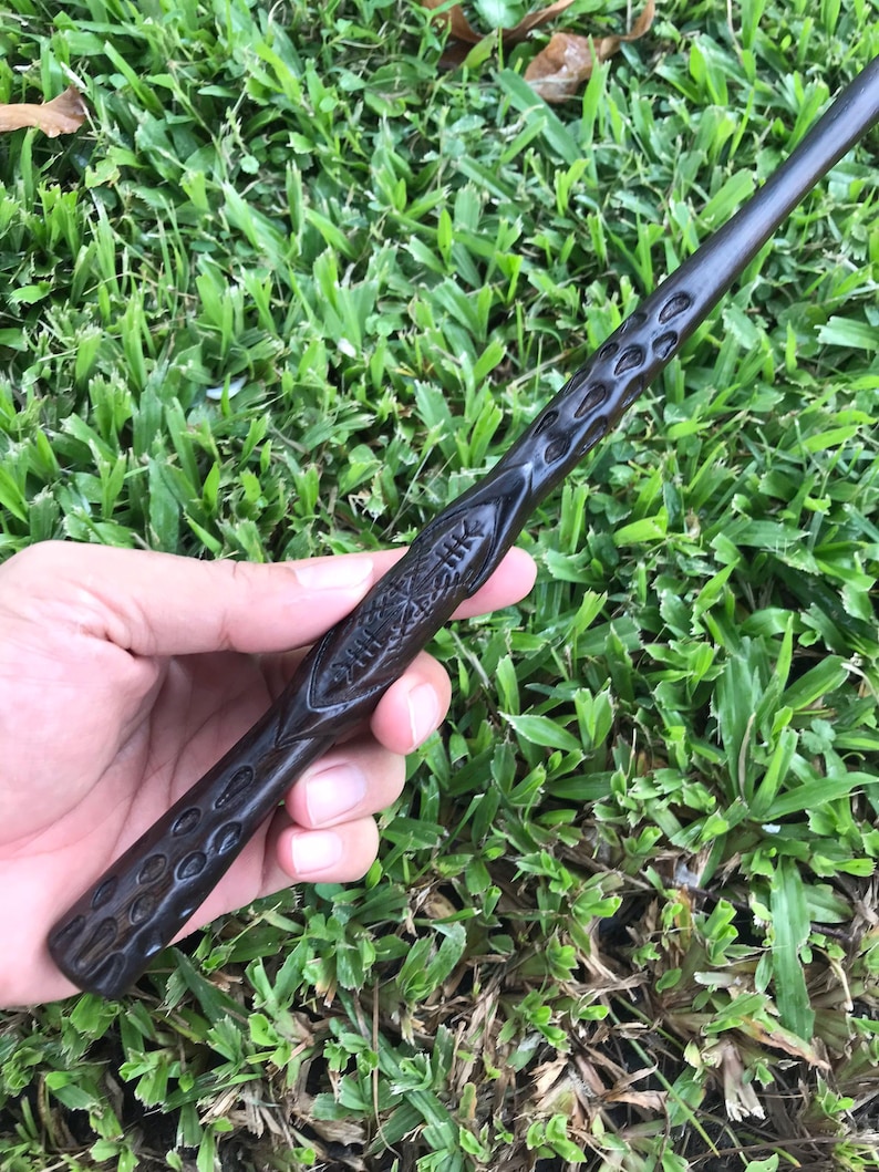 May include: A dark brown wooden wand with intricate carvings. The wand is held in a hand, with the tip of the wand pointing towards the right.