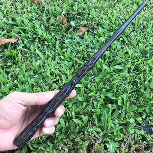 May include: A dark brown wooden wand with intricate carvings. The wand is held in a hand against a green grassy background.