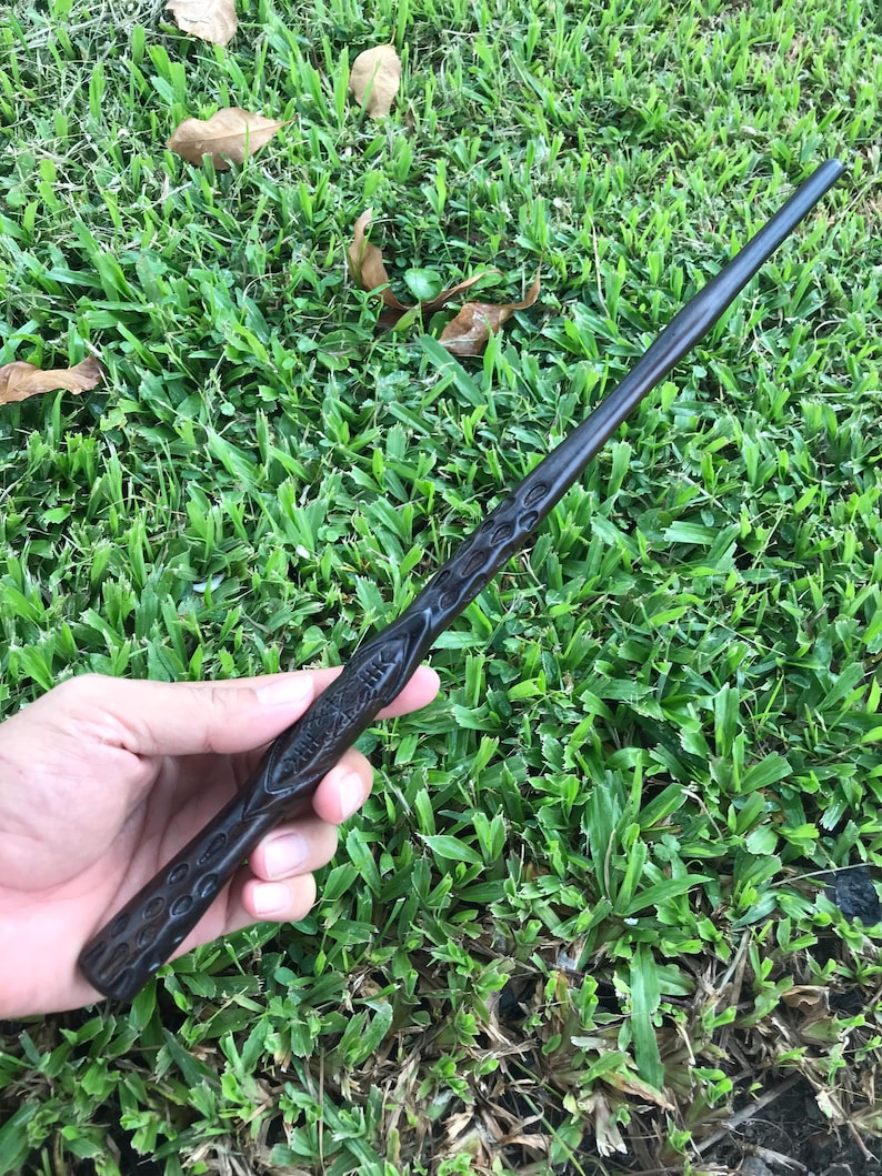 May include: A dark brown wooden wand with intricate carvings. The wand is held in a hand against a green grassy background.