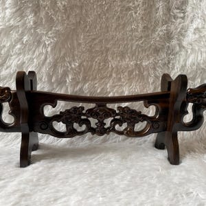 Puede incluir: Un soporte para espada de madera marrón oscuro con intrincados tallados de dragones y diseños florales. El soporte tiene dos patas y está diseñado para sostener una espada u otra hoja larga.