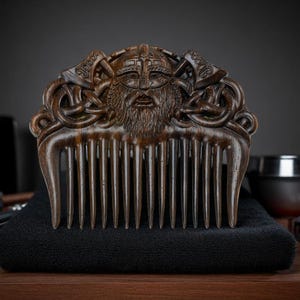 May include: A dark brown wooden comb with a detailed carving of a Viking warrior's face and helmet. The comb features a wide handle with intricate knotwork and two axes. The comb is resting on a black towel.