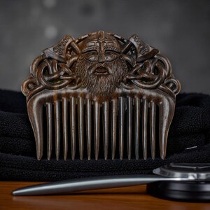 May include: A wooden comb with a carved Viking design featuring a bearded face, helmet, and axes. The comb is dark brown and rests on a black towel, with a silver grooming tool below.