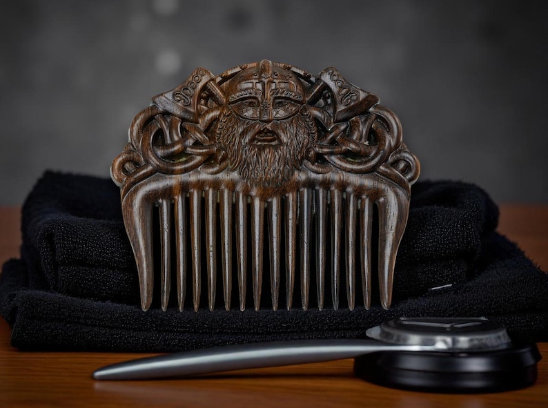May include: A wooden comb with a carved Viking design featuring a bearded face, helmet, and axes. The comb is dark brown and rests on a black towel, with a silver grooming tool below.