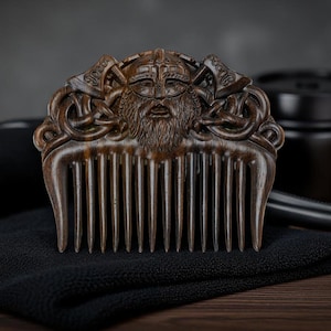 May include: A wooden comb with a detailed carving of a Viking warrior's face, complete with a helmet and beard. The comb is brown and rests on a black cloth, with other grooming items in the background.
