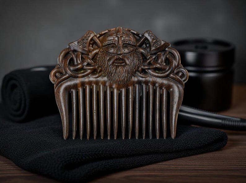 May include: A wooden comb with a detailed carving of a Viking warrior's face, complete with a helmet and beard. The comb is brown and rests on a black cloth, with other grooming items in the background.