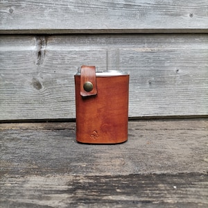 Peut inclure: Un porte-flacon en cuir marron avec un flacon en argent et une fermeture à pression en métal. Le porte-flacon a un petit logo sur le devant.