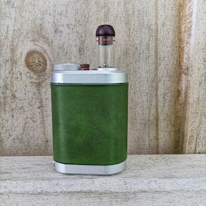 Peut inclure: Un appareil vert recouvert de cuir, avec des garnitures argentées. Il comporte un tube en verre transparent avec un dessus en bois brun et une bande rouge. L'appareil est rectangulaire, avec une petite pièce en bois ronde sur le dessus.