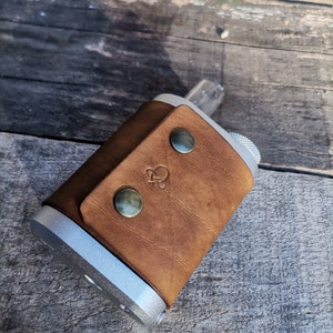Peut inclure: Un étui en cuir marron avec un appareil argenté à l'intérieur. L'étui a trois boutons-pression métalliques et un logo sur le devant.