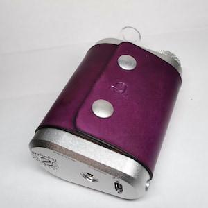 Peut inclure: Un étui en cuir violet avec des détails argentés et deux fermetures à boutons argentés. L'étui entoure un appareil argenté avec un dessus texturé et un embout buccal en verre transparent. L'appareil porte le texte "TinyMight2" en bas.
