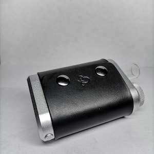 Peut inclure: Un vaporisateur noir et argenté avec un embout buccal en verre transparent. L'appareil a une forme rectangulaire avec des bords arrondis et deux boutons argentés sur le dessus. L'appareil a une finition noire texturée semblable à du cuir.