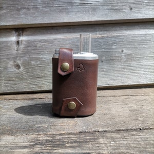 Peut inclure: Un étui en cuir marron avec des fermetures à boutons en laiton, contenant un appareil en argent et en verre. L'étui présente un petit motif en relief et enveloppe l'appareil. Le fond est constitué de planches de bois patinées.