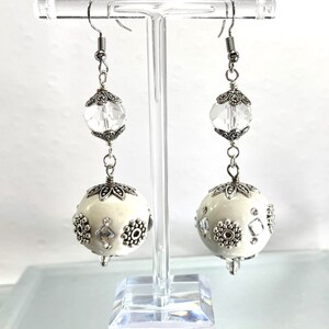 May include: A pair of long, silver dangle earrings with white, round, beaded ornaments. The ornaments are decorated with silver accents and clear rhinestones.