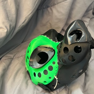 May include: A black and green dog mask with a mesh design and a large eye hole. The mask is made of a hard plastic material and has a strap that goes around the head.