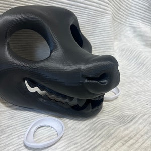 Könnte beinhalten: Eine schwarze, im 3D-Druck hergestellte Tiermaske mit offenen Augenlöchern und einem zahnigen Grinsen. Die Maske besteht aus Kunststoff und hat eine strukturierte Oberfläche. Zwei weiße Kunststoff-Augeninserts sind im Lieferumfang enthalten.