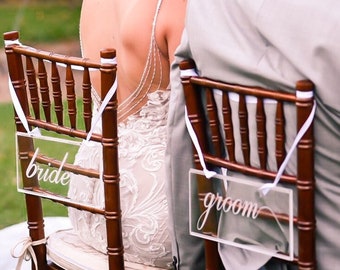 May include: A bride and groom are seated at a table with a floral centrepiece. The bride is wearing a white lace dress and the groom is wearing a grey suit. The chairs are decorated with signs that say "bride" and "groom".