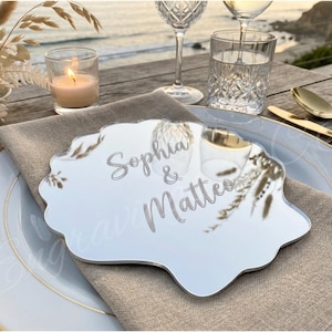 May include: A mirrored place card with the names "Sophia & Matteo" engraved on it, set on a table with a linen napkin, plate, and gold cutlery. A lit candle, glassware, and decorative flowers complete the elegant table setting.