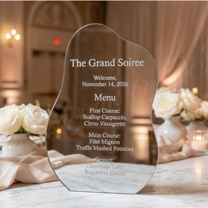 May include: A clear, uniquely shaped menu card for "The Grand Soiree" with engraved text. The menu lists courses like Scallop Carpaccio and Filet Mignon. The date is November 14, 2026. White roses and candles are in the background.