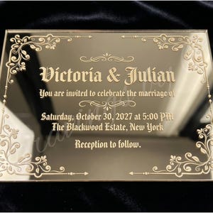 May include: A rectangular, gold-colored acrylic wedding invitation. The text reads "Victoria & Julian" in an elegant font, with the wedding date, time, and location. Decorative floral corner accents frame the text, with the words "Reception to follow."