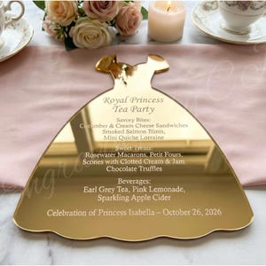 May include: A gold-colored acrylic dress-shaped menu for a Royal Princess Tea Party. The menu lists savory bites, sweet treats, and beverages. The event is a celebration for Princess Isabella on October 26, 2026.