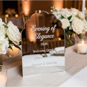 May include: A mirrored sign with a scalloped top, displaying the text "Evening of Elegance 2026" and event details. The sign is on a table with white floral arrangements and lit candles, creating an elegant ambiance.
