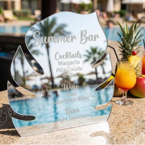 May include: A clear acrylic Summer Bar sign in a leaf shape, reflecting a pool and palm trees. The sign lists cocktails, wine, and beer options. Tropical drinks and fruit are arranged on a countertop.