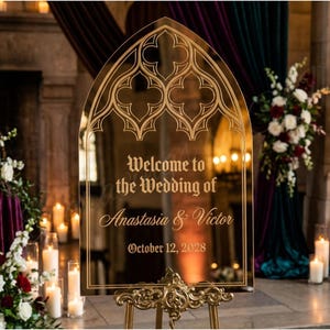 May include: A gold-framed mirror sign with an arched top, displaying the text "Welcome to the Wedding of Anastasia & Victor, October 12, 2028." The sign is on a decorative gold stand, surrounded by flowers and candles, creating a romantic wedding atmosphere.