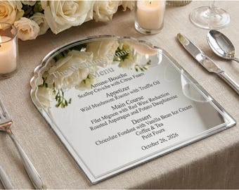 May include: A mirrored wedding dinner menu with a scalloped top edge, displaying a detailed menu and the date October 26, 2026. The menu is on a table with cutlery, candles, and floral arrangements.
