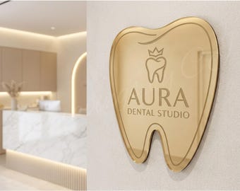 May include: A gold-coloured sign in the shape of a tooth, with the words "AURA DENTAL STUDIO" in a light font. The sign features a tooth and crown graphic. The background is a dental office interior.