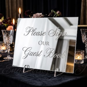 May include: A mirrored sign with the text "Please Sign Our Guest Book" in elegant script. The sign is displayed on a table with crystal glassware, candles, and floral arrangements, creating a sophisticated ambiance.