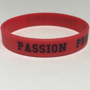 May include: A red silicone wristband with the words "PASSION PRO" printed in black block letters. The band is a solid, vibrant red color and appears to be flexible. The text is centered on the band.