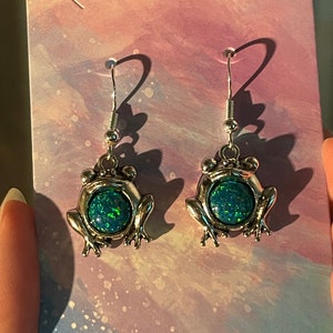 May include: A pair of silver frog earrings with green opal stones. The earrings have a dangling design with a hook closure.