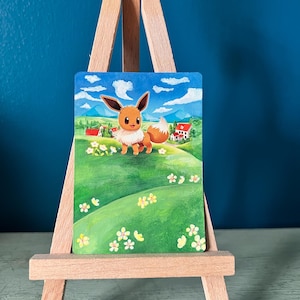 May include: A rectangular painting featuring a brown Eevee character standing in a vibrant green meadow filled with small yellow flowers. The background displays a blue sky with white clouds, distant mountains, and small houses on a wooden easel.