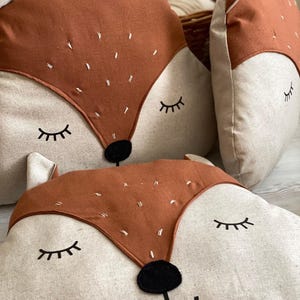 May include: Two brown and beige fox-shaped pillows with embroidered eyes and noses. The pillows are made of a soft, plush fabric and are perfect for adding a touch of whimsy to any room.