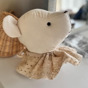Può includere: Un peluche di stoffa bianca a forma di topo con un occhio marrone e un colletto di pizzo beige.