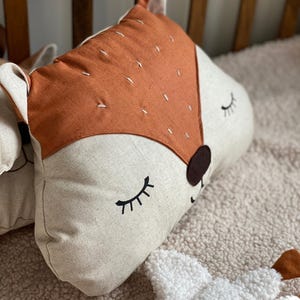 May include: A plush pillow shaped like a fox with a brown and white face and a tan nose. The fox has closed eyes with black eyelashes and a stitched smile. The pillow is lying on a white fluffy surface.