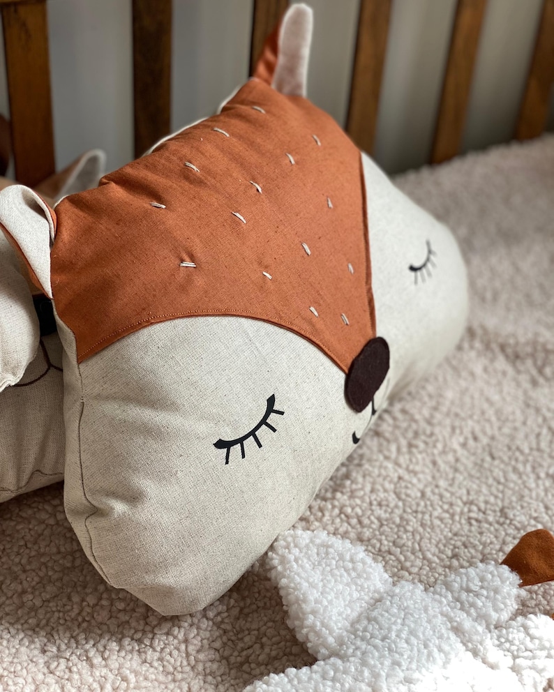 May include: A plush pillow shaped like a fox with a brown and white face and a tan nose. The fox has closed eyes with black eyelashes and a stitched smile. The pillow is lying on a white fluffy surface.
