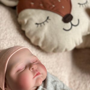 May include: A sleeping baby doll wearing a pink hat and a gray onesie with a button. The doll is lying on a soft, fuzzy blanket next to a brown and white deer-shaped pillow.
